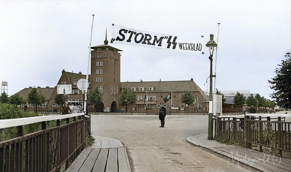 In ons archief vonden wij deze zwart-wit foto genomen door de NSB fotodienst. Op zogenaamde contactafdrukken. Deze foto hebben we ingekleurd. Deze foto maakte dat we meer wilde weten over de achtergrond. De foto is genomen bij de Veemarkt in Den Bosch. Het is vrij uniek omdat men nog over een bruggetje moest om het terrein te betreden. Op de foto is een spandoek te zien met de tekst; “Storm SS weekblad”. Het is een spandoek dat de propaganda uiting goed laat zien. Het tijdschrift “Storm SS” werd uitgegeven door de Nederlandse SS om publiek geïnteresseerd te krijgen en lid te laten worden in de gedachtengoed van de SS.